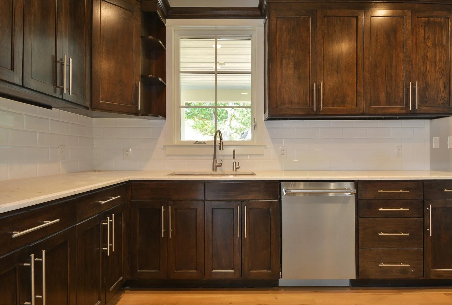 Kitchen with dark wooden cabinets, white countertop, white backsplash, window above sink, and built-in dishwasher.