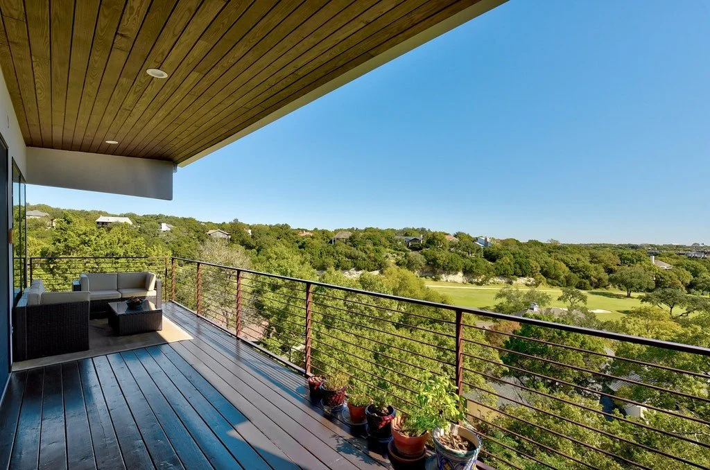 Balcony with outdoor furniture overlooking a green landscape, potted plants along the railing, under a wood-paneled ceiling.