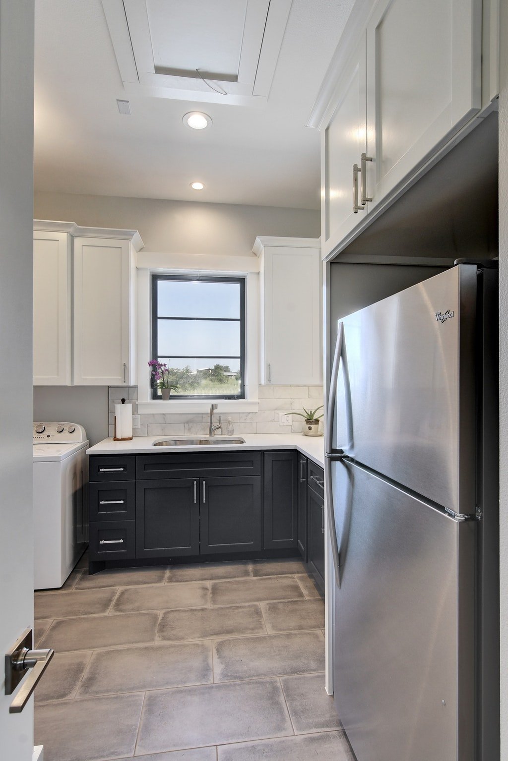 Modern kitchen with white and black cabinetry, stainless steel refrigerator, window with a potted plant, and tiled floor.