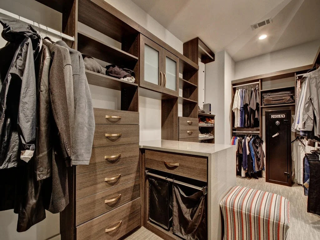 Walk-in closet with clothes hanging on rods, wooden shelves and drawers, a safe, and a small striped ottoman.