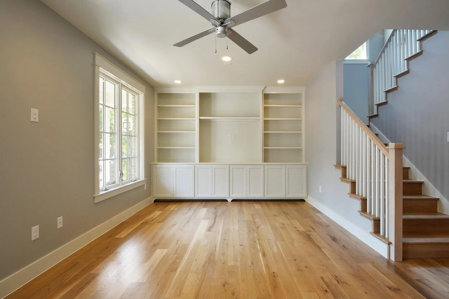 Empty living room with large window, built-in white shelves, hardwood floors, and staircase with wooden handrail.