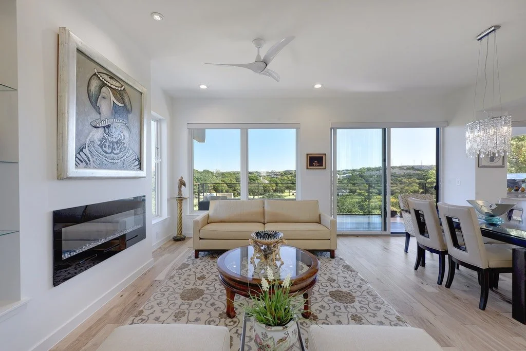 Modern living room with white walls, cream sofa, glass-top coffee table, large windows with a view of trees, indoor planted flower, chandelier, dining area with white chairs, and artwork on the wall.