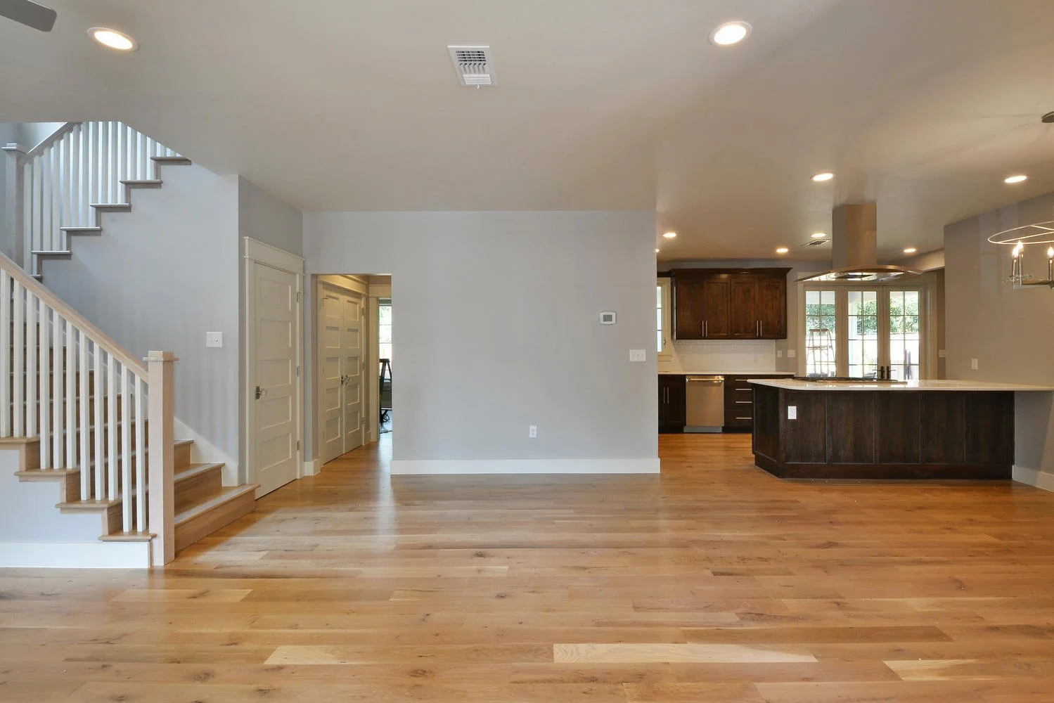 Empty living room with hardwood floors, light gray walls, staircase on the left, open kitchen with dark wood cabinets and white countertops, large windows, and recessed ceiling lights.