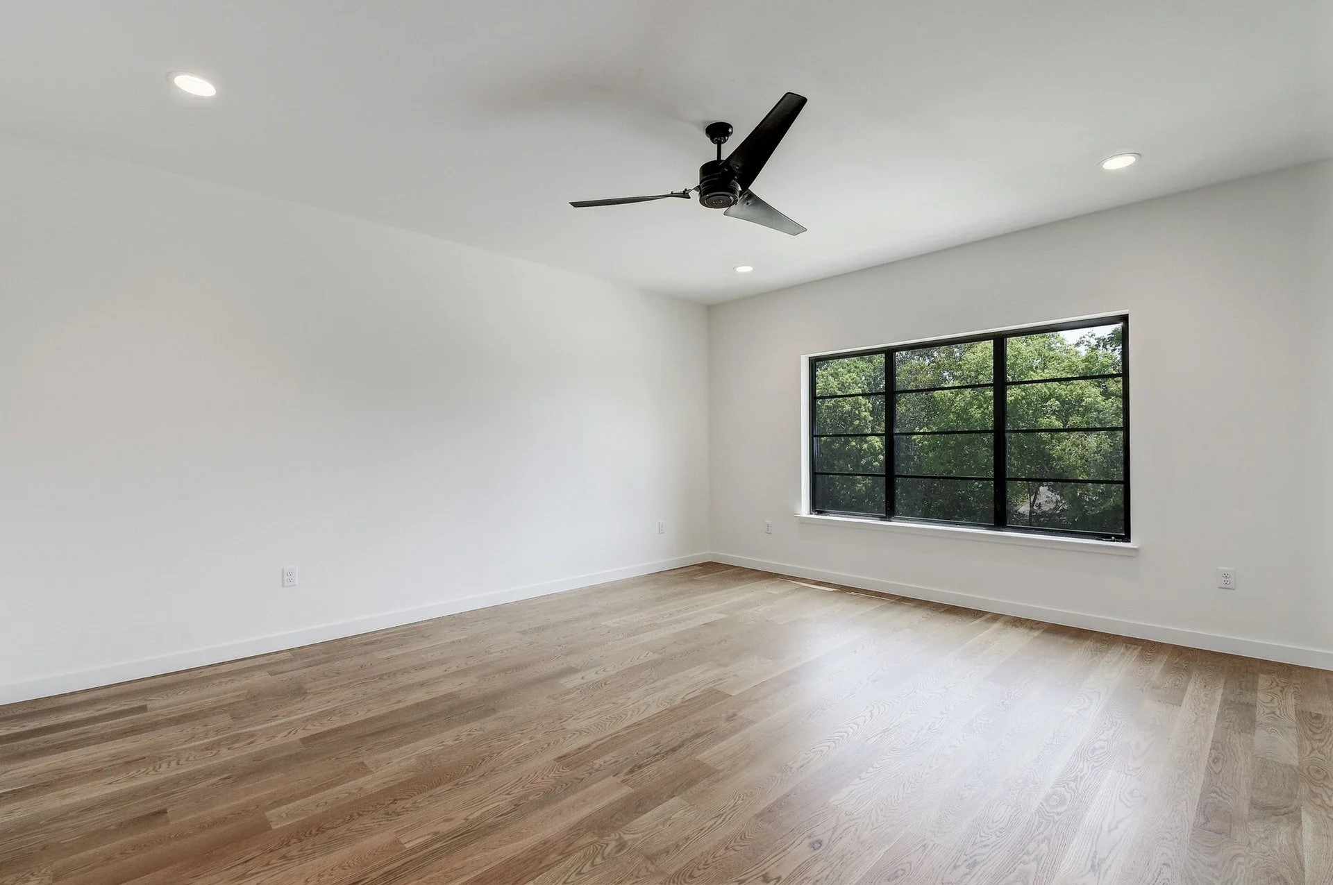 Empty room with white walls, wood flooring, large window, black ceiling fan, and recessed lighting.