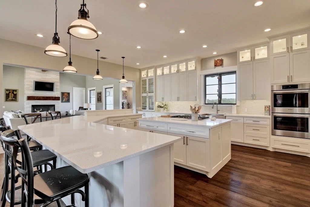 Modern open-concept kitchen with white cabinets, a large island with a white countertop, and dark wood flooring. There are pendant lights above the island, a sink under a window, and double ovens built into the cabinetry. In the background, a living 