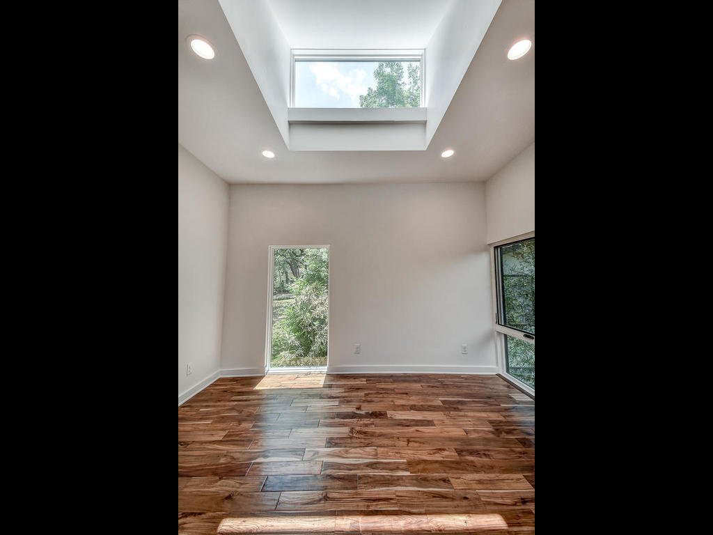 Empty room with white walls, hardwood flooring, a skylight, a window, and a door leading outside with trees visible outside.
