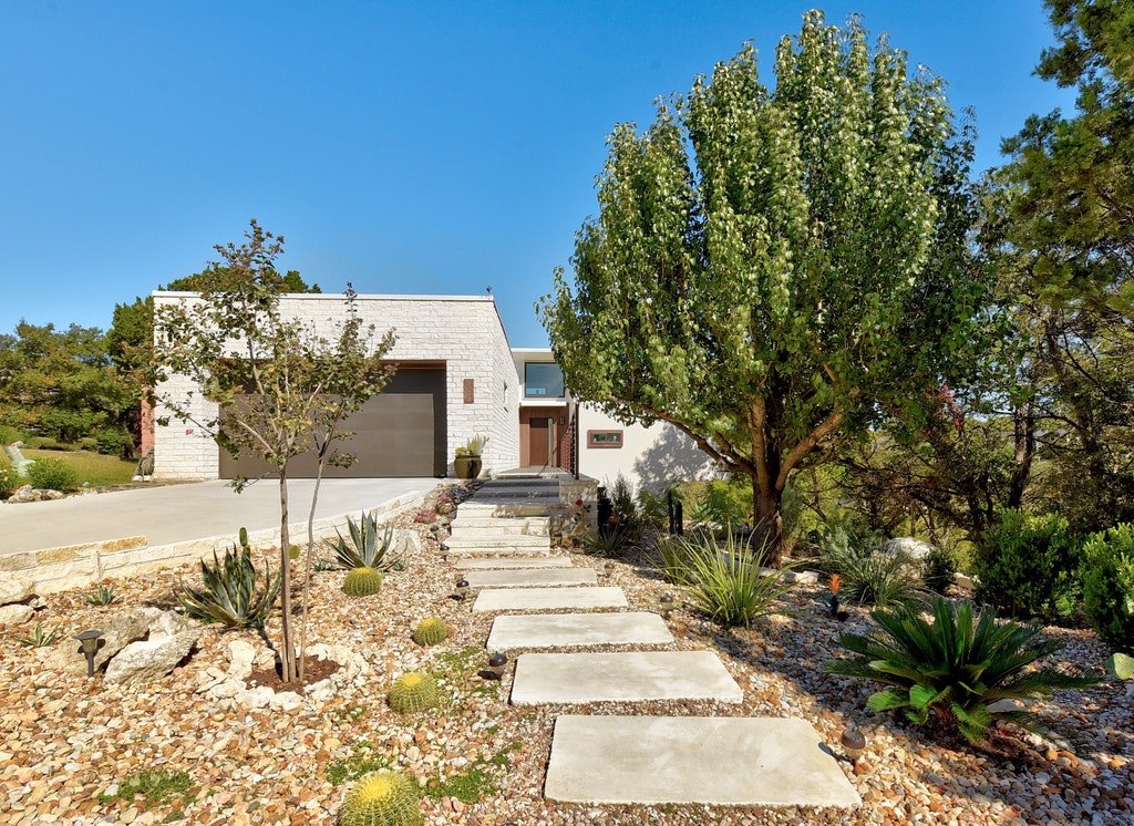 Modern white house with a two-car garage, surrounded by desert landscaping, including cacti, agave plants, and trees, under a clear blue sky.