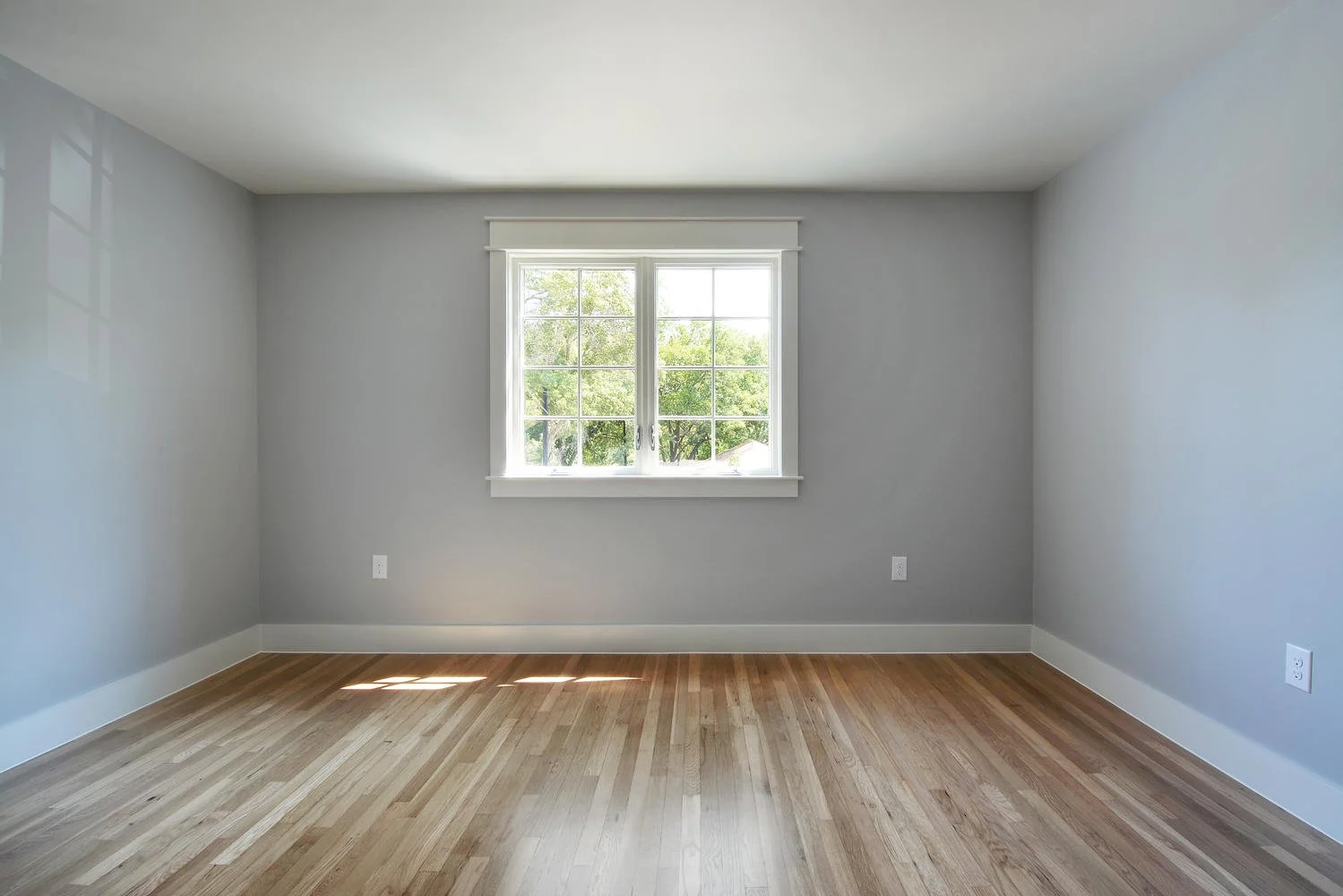 Empty room with light gray walls, a window with white trim, and hardwood flooring.