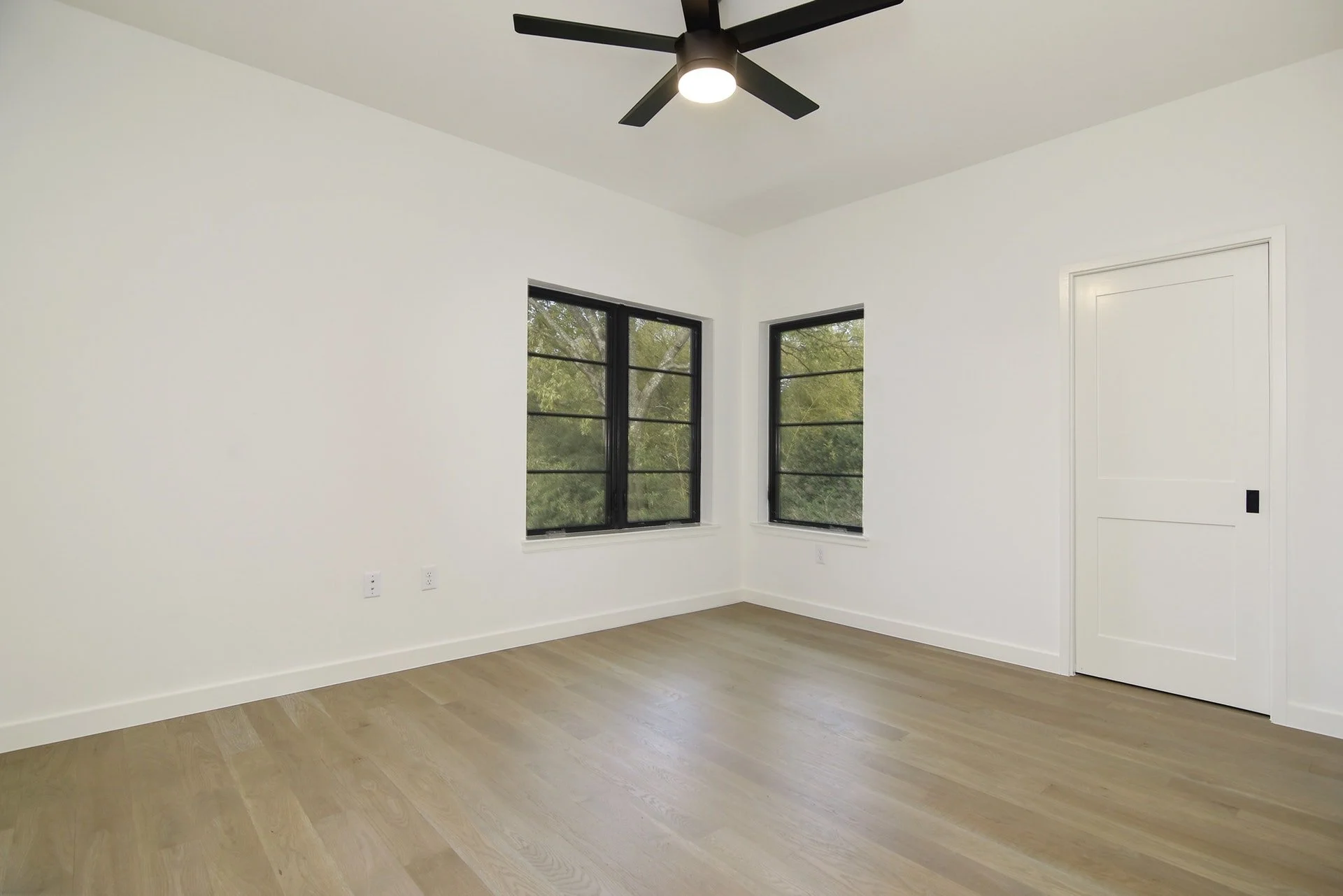 Empty room with white walls, two black-framed windows showing green trees outside, a white door, a modern black ceiling fan with light, wood flooring, and electrical outlets.