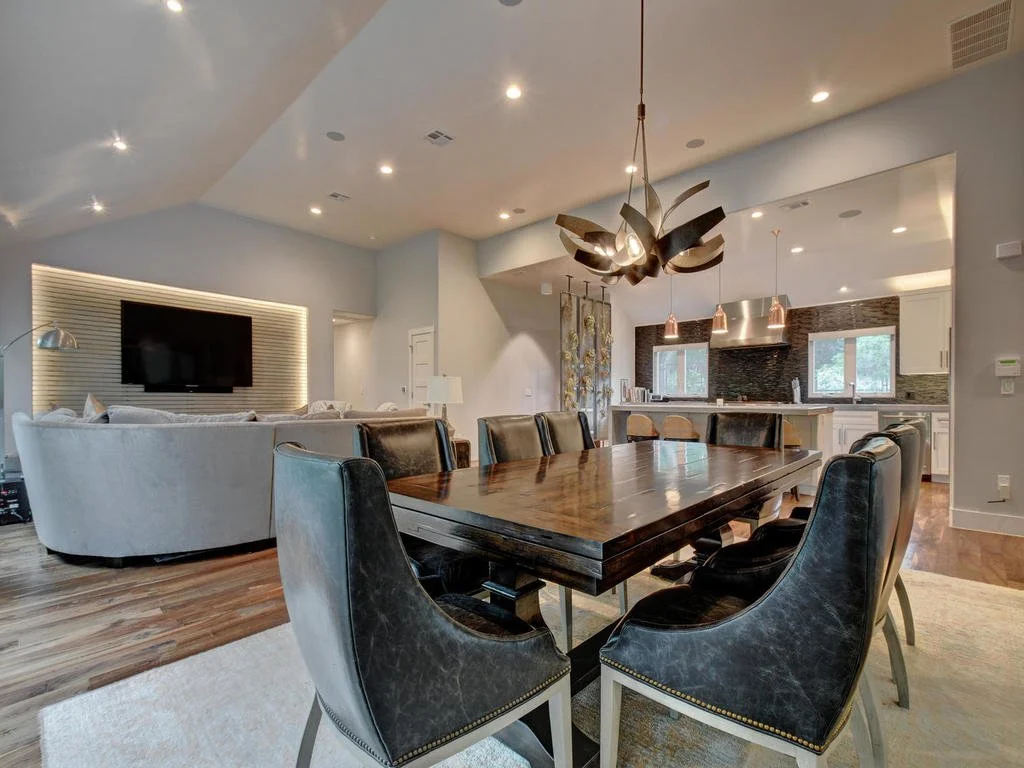 Open-concept living and dining area with a large wooden dining table and black leather chairs, modern chandelier, and a living room with a curved white sectional sofa facing a wall-mounted TV. Kitchen in the background with pendant lighting, windows,