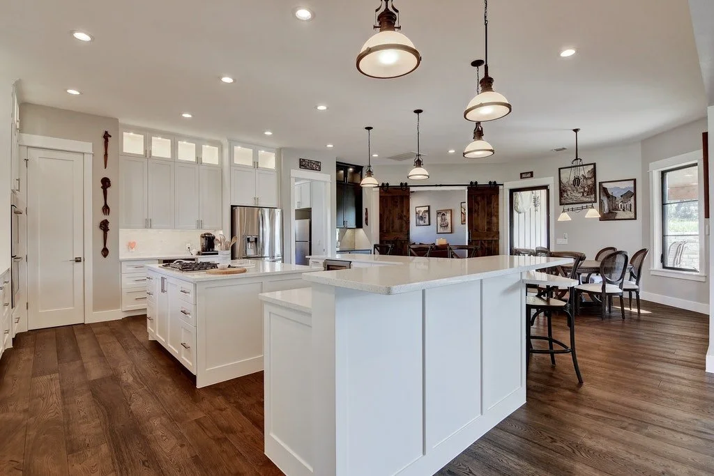 Modern kitchen with white cabinets, a large island, stainless steel appliances, wooden floors, hanging pendant lights, and a dining area with chairs and framed pictures.