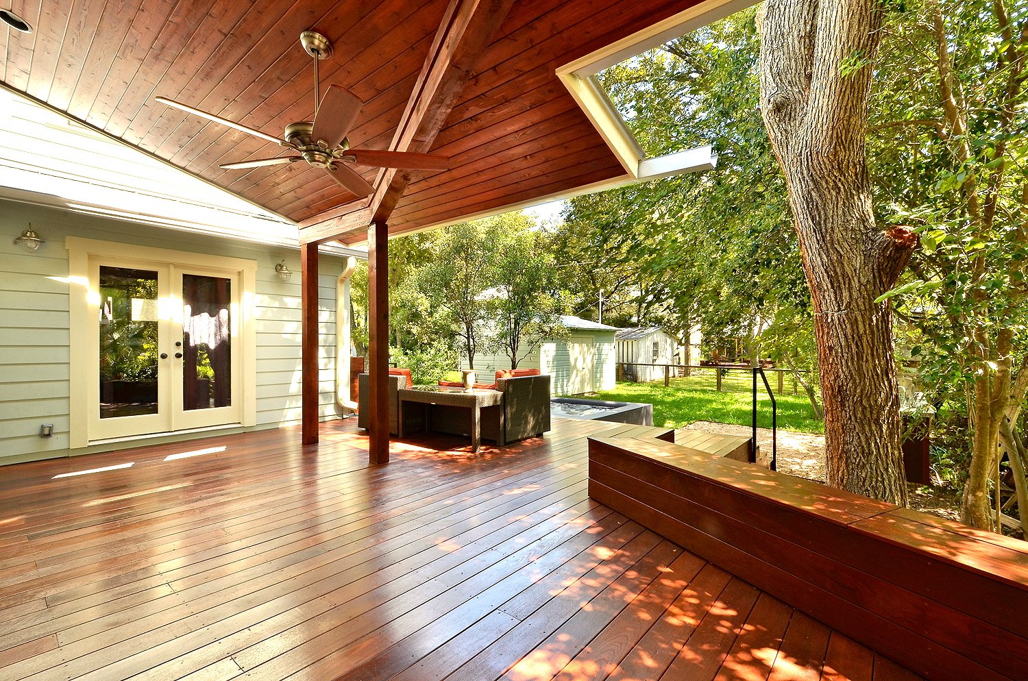 A spacious outdoor wooden deck attached to a house, with a ceiling fan, patio furniture, and surrounded by trees and greenery in a backyard.