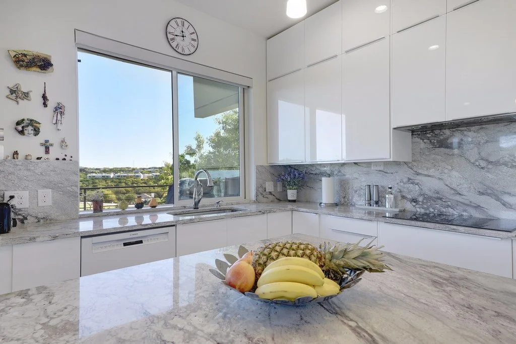 Modern kitchen with white cabinets, marble countertops, and a bowl of bananas, pineapple, and apple on the island. There is a large window with a view of trees and a balcony, a wall clock, and various small decorative items on the counter.
