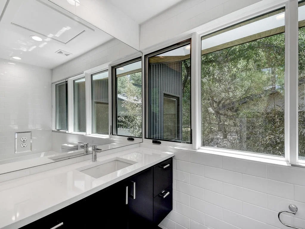 Modern bathroom with white tiled walls and a large mirror. Multiple windows reveal green trees outside, and there is a white countertop with a built-in sink and a black cabinet below.