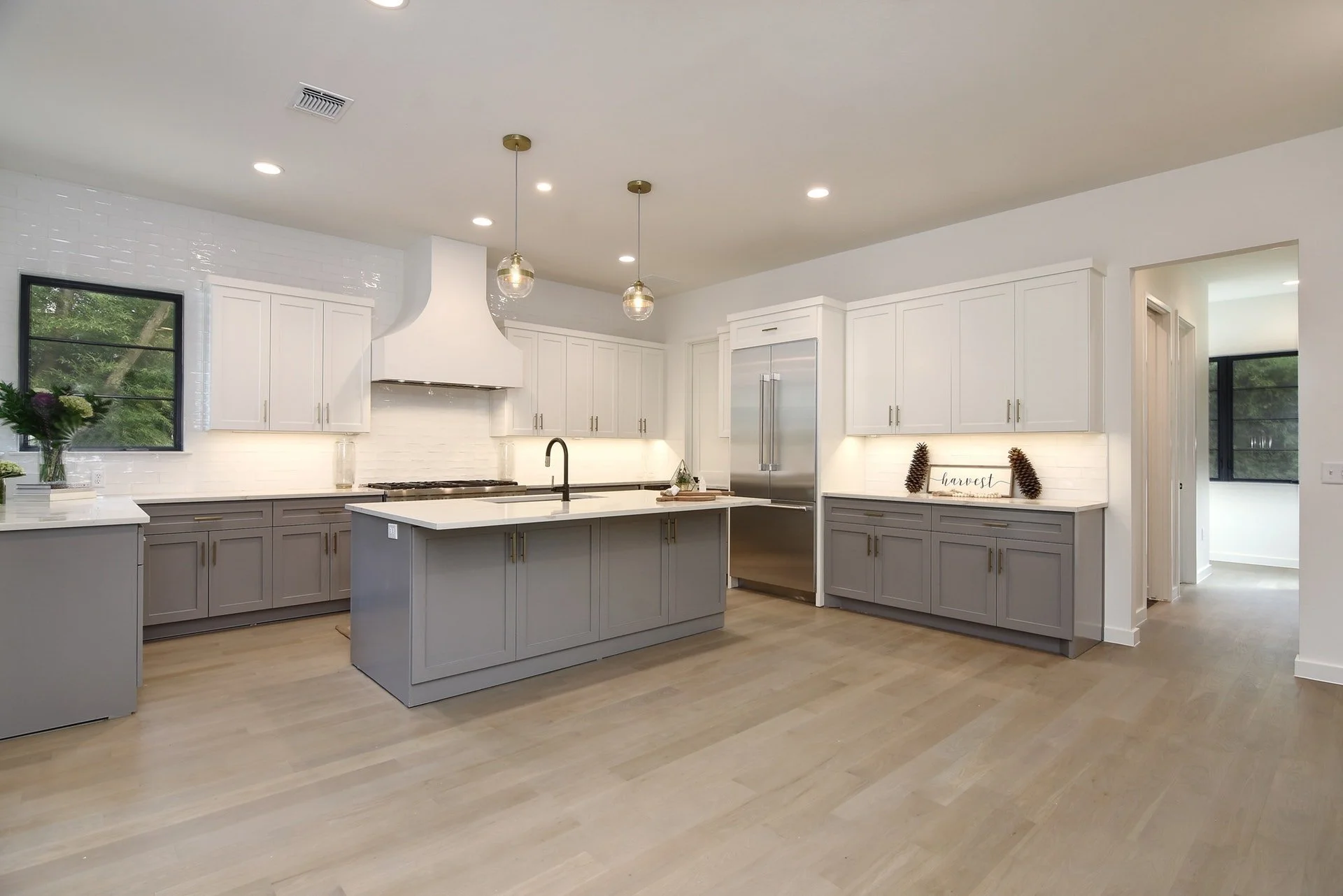 Modern white and gray kitchen with two pendant lights, large island with sink, stainless steel refrigerator, and decor including pinecones and a wooden sign that reads 'harvest'.