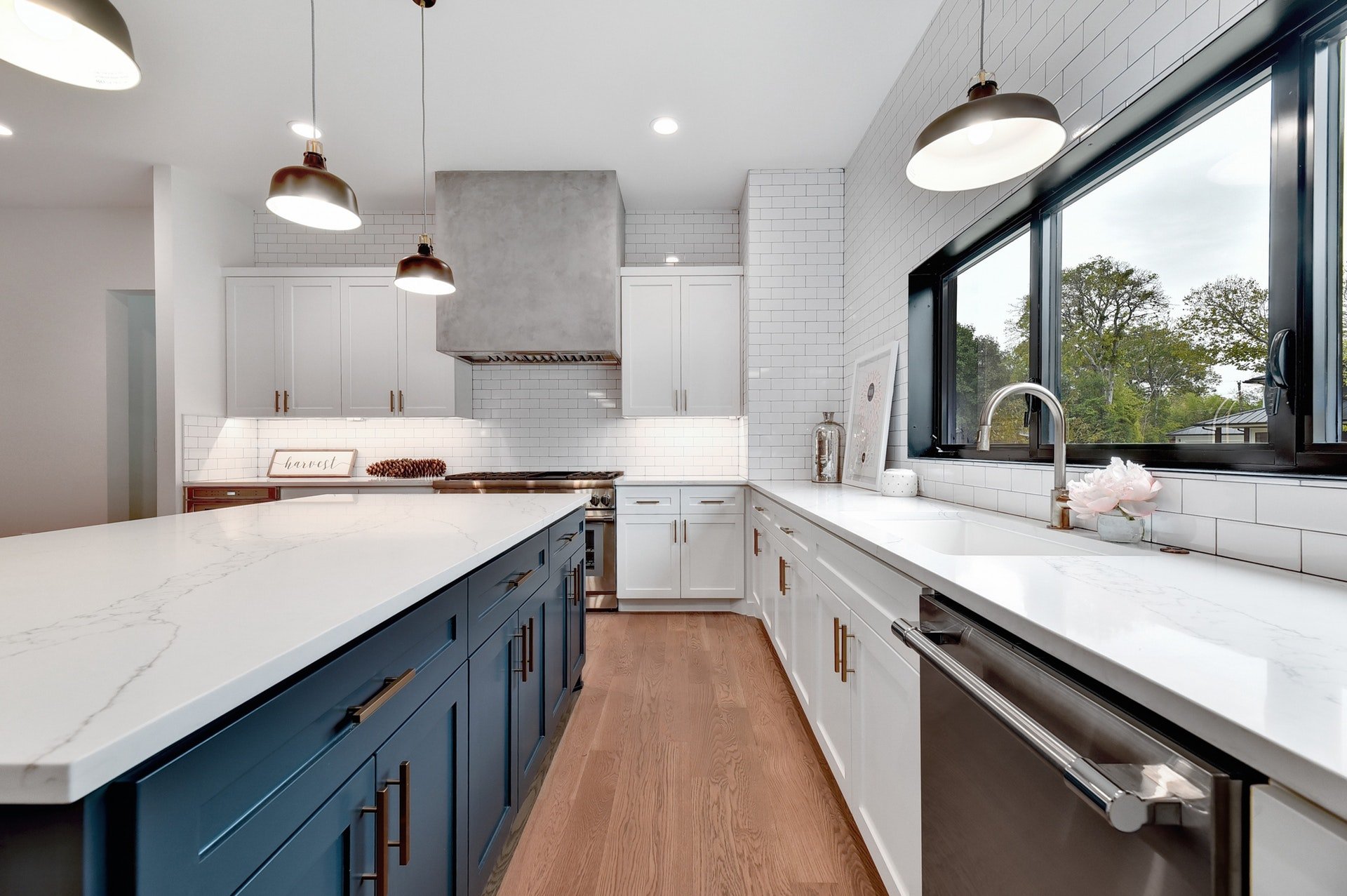 Modern kitchen with white cabinets, a large white marble countertop island, blue lower cabinets, and wood flooring. There are three pendant lights hanging from the ceiling, a stainless steel range hood, and a large window above the sink overlooking t