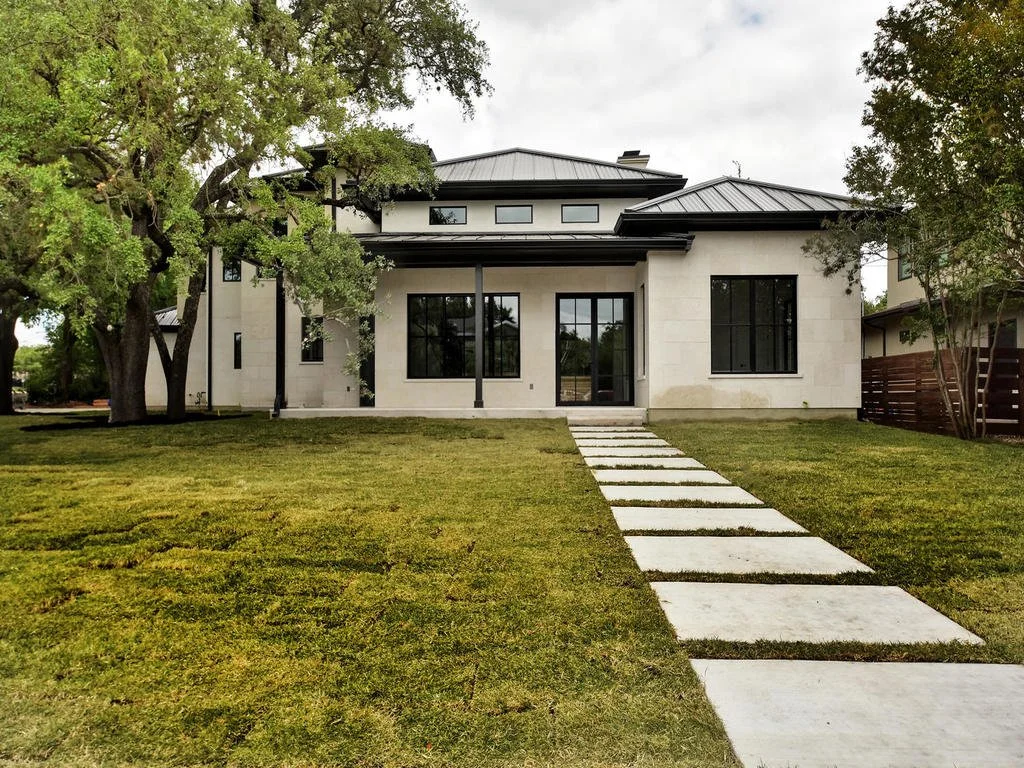 Modern white house with black window frames and a metal roof, surrounded by green lawn and trees, with a concrete walkway leading to the front entrance.