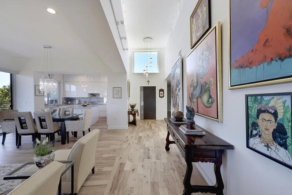 Living area with a hallway view, showcasing a gallery wall of colorful framed paintings and a dark wooden console table with various sculptures and books, hardwood floors, and a partially visible kitchen with a dining area and bar stools.