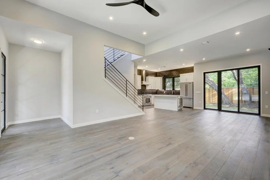 Open-concept living space with hardwood floors, a staircase leading upstairs, a modern kitchen with white cabinets and stainless steel appliances, and large sliding glass doors opening to a backyard with trees.