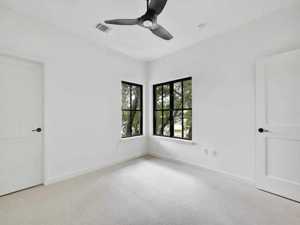 Empty white room with two black-framed windows showing trees outside, two white doors with black handles, a black ceiling fan, white walls, and off-white carpet.