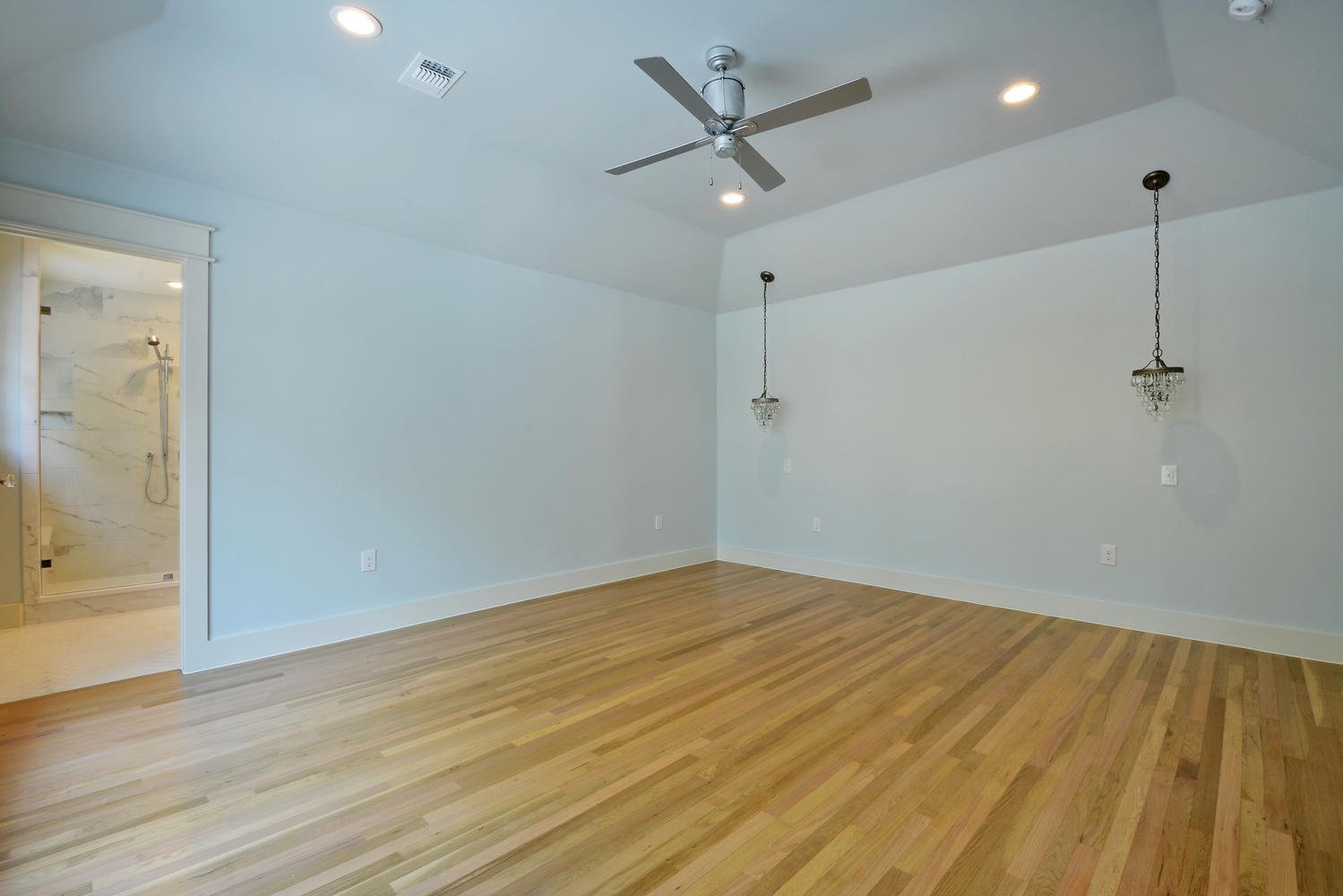 Empty living room with hardwood floors, two hanging light fixtures, ceiling fan, and visible bathroom with shower in the background.