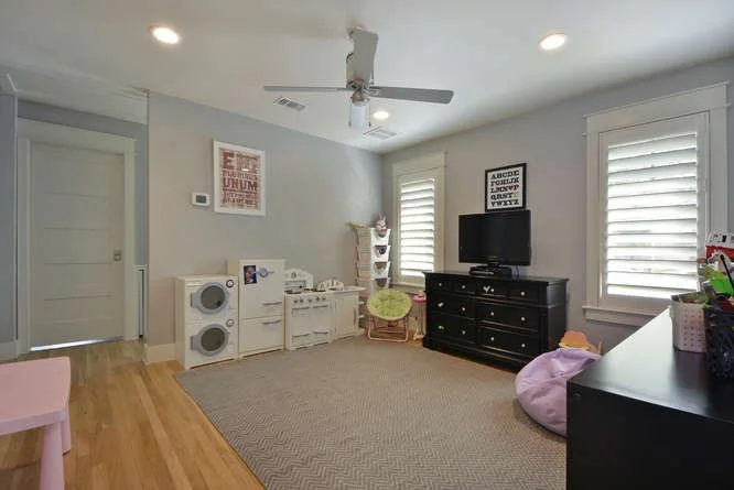 Living room with light gray walls, hardwood floors, a ceiling fan, and two windows with white shutters. Contains a black TV stand with a flat-screen TV, a white laundry or storage unit with laundry baskets and a small desk on top. There's a green cha
