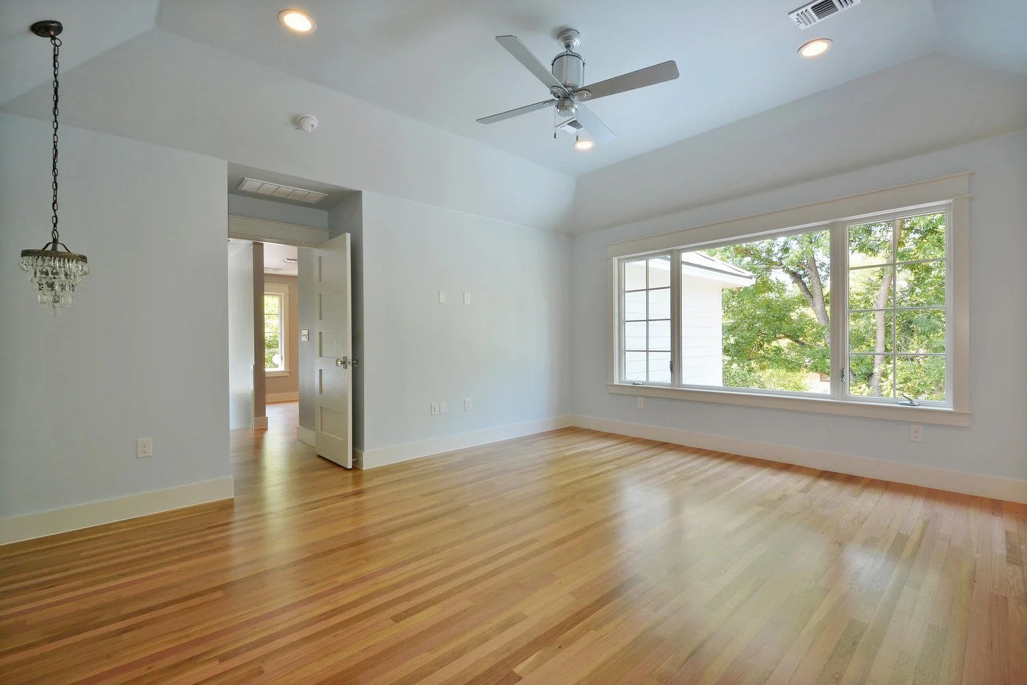 Empty living room with large window, hardwood floors, white walls, ceiling fan, and recessed lighting.