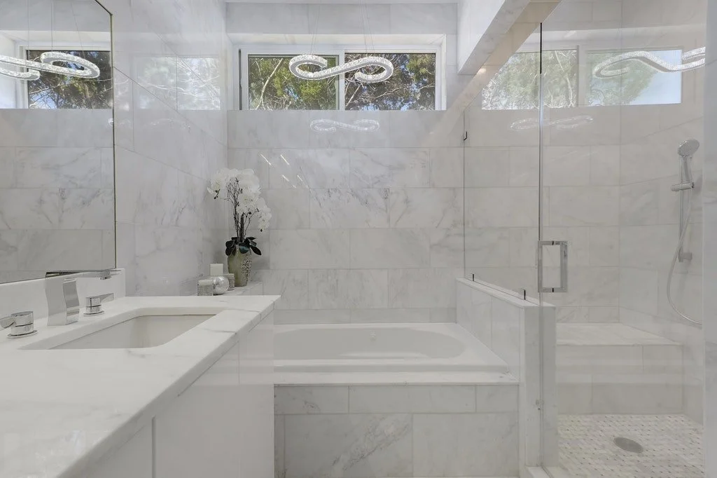 Modern bathroom with white marble walls, bathtub, and a walk-in shower, decorated with a potted orchid plant and candles.