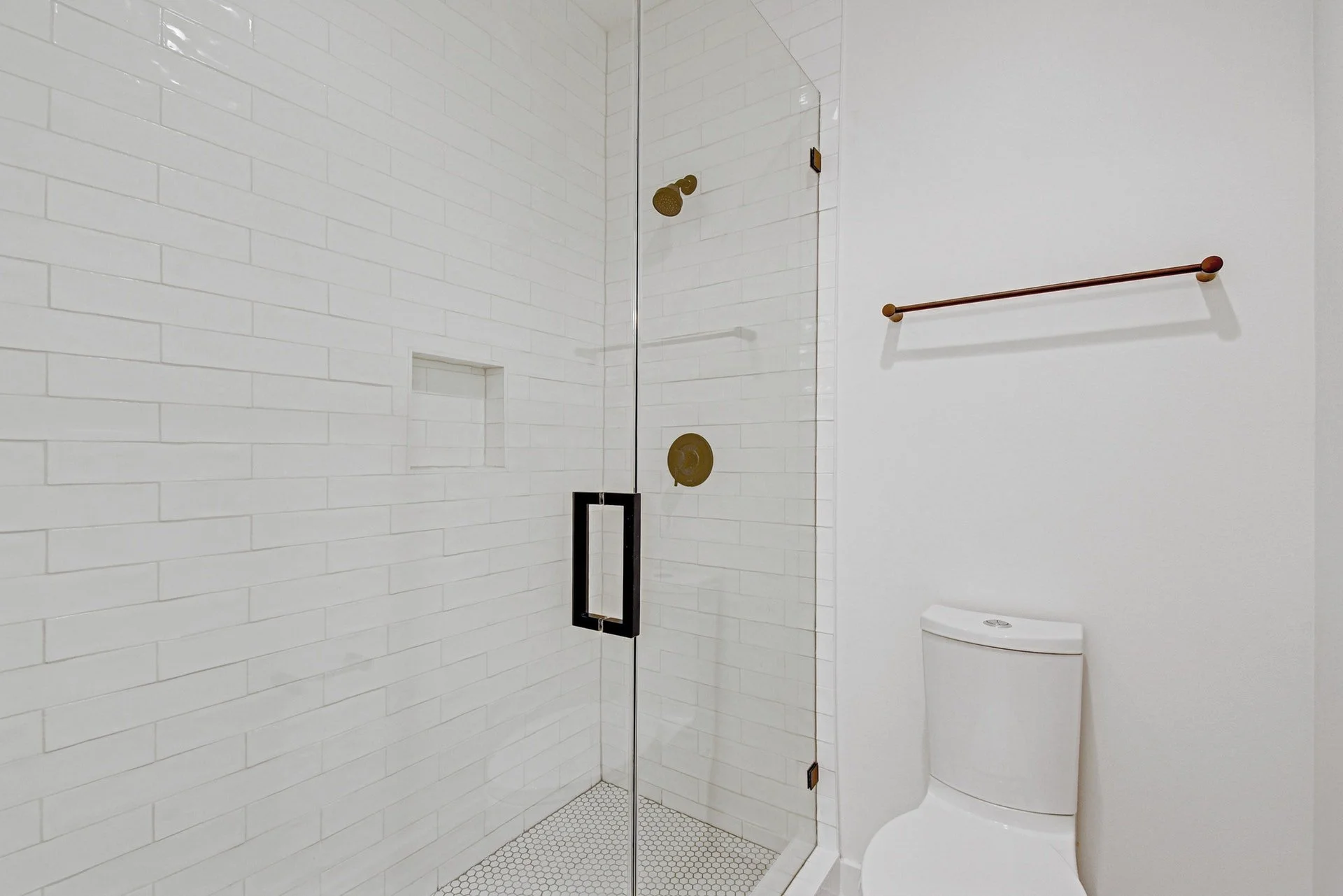 A modern bathroom with a walk-in shower featuring white subway tiles, a black-framed glass door, and a built-in niche. Next to the shower is a white toilet and a simple wooden towel bar mounted on the wall.