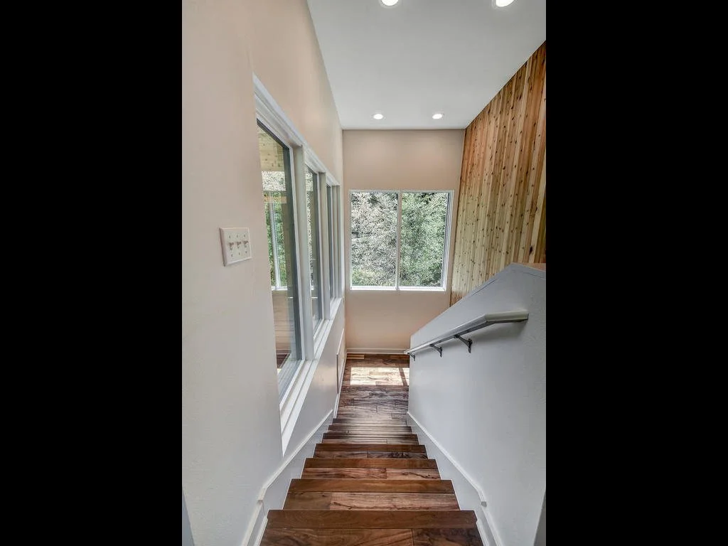Wooden staircase inside a house with white walls, large windows, a wood-paneled accent wall, and natural light.