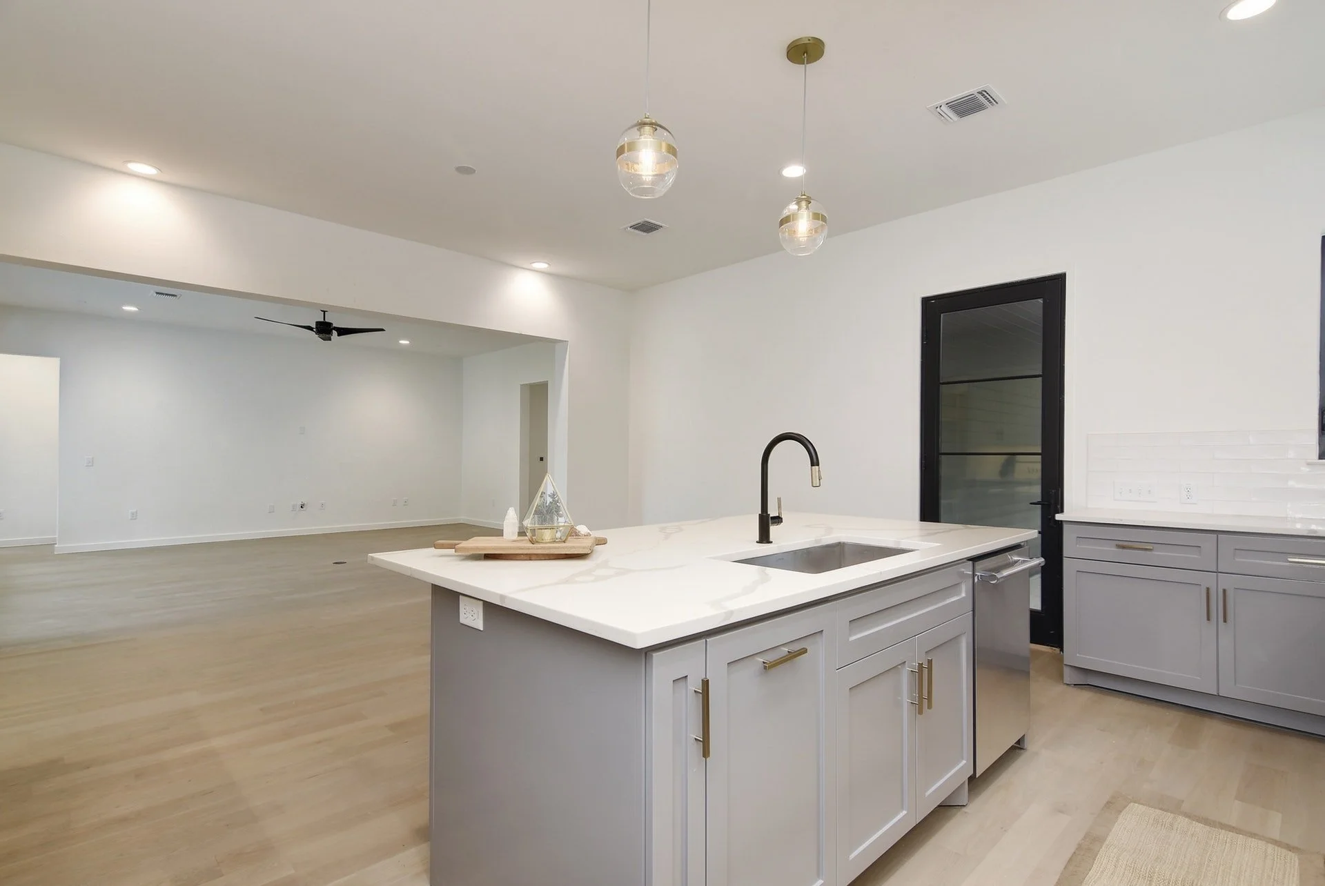 Open-concept kitchen and living space with white walls, light hardwood floors, modern pendant lighting, a black ceiling fan, and a large island with a sink and gray cabinets.