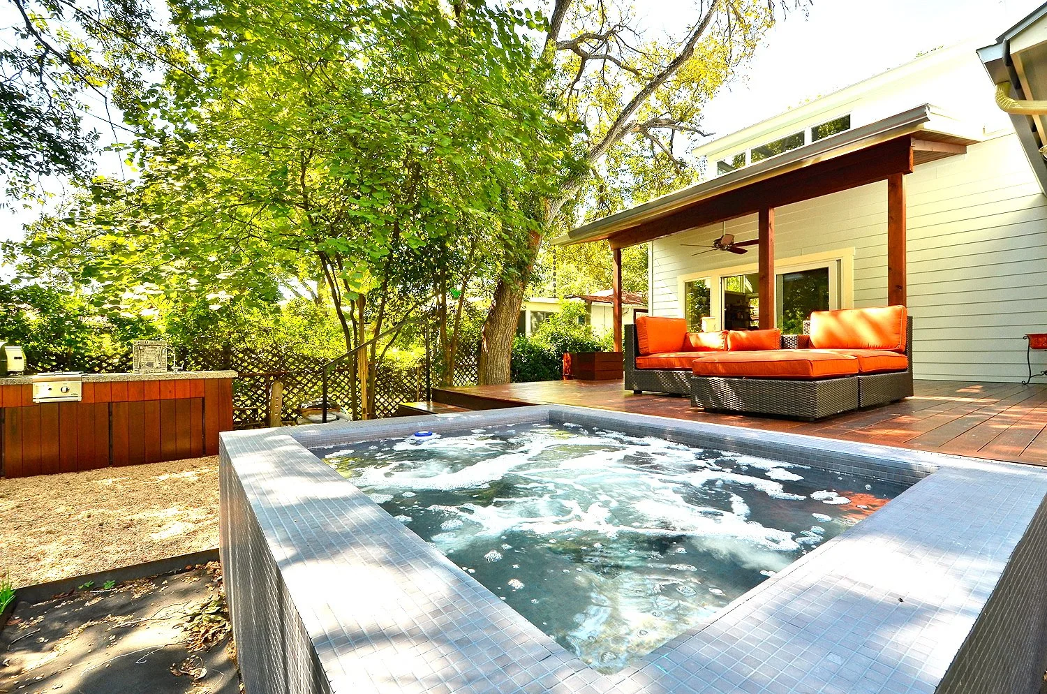Backyard patio with hot tub, outdoor seating with orange cushions, deck area, and surrounding trees in a sunny day.