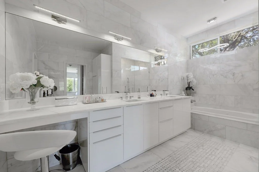 A modern bathroom with white marble walls, a large mirror, and a white vanity with multiple drawers. The vanity has two sinks and is decorated with flowers and toiletries. There is a bathtub to the right with a white towel hanging nearby. Natural lig