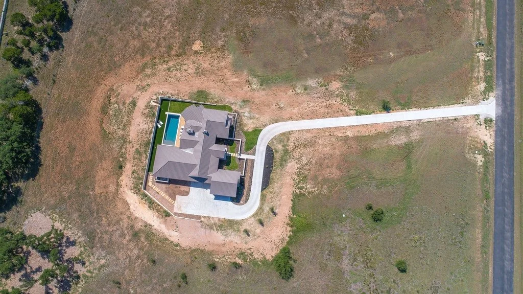 An aerial view of a house with a driveway, pool, and surrounding open land.