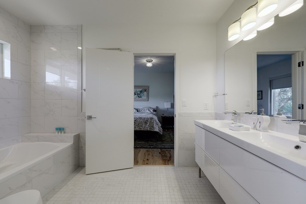 Modern bathroom with white marble walls, a large mirror, a white vanity with double sinks, and a window. The bathroom door is open, revealing a bedroom with a bed and a nightstand.