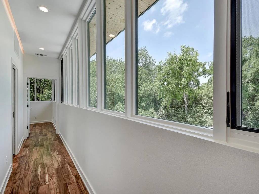 A bright enclosed porch with large windows overlooking trees, white walls, and wooden flooring.