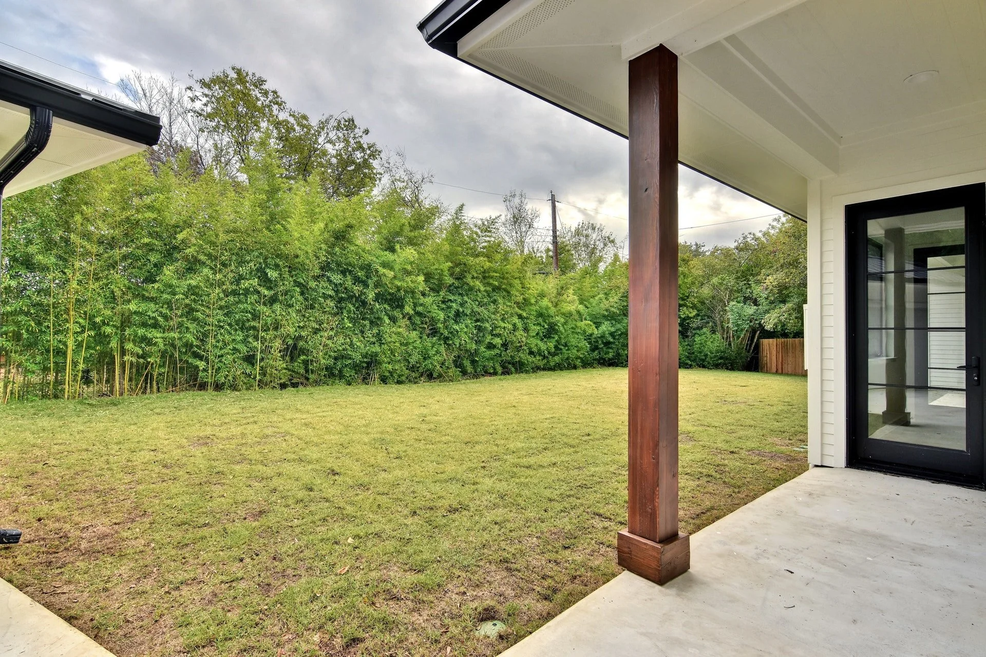 View of a backyard with a grassy lawn, surrounded by green trees and shrubbery, seen from a covered patio area with a concrete floor, a wooden support beam, and a nearby modern house with a glass door.