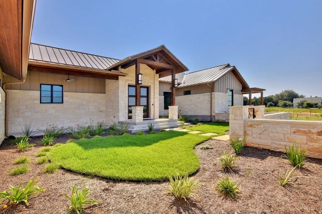 Modern house with stone walls, metal roof, and a front porch, surrounded by a landscaped yard with green grass and small plants.