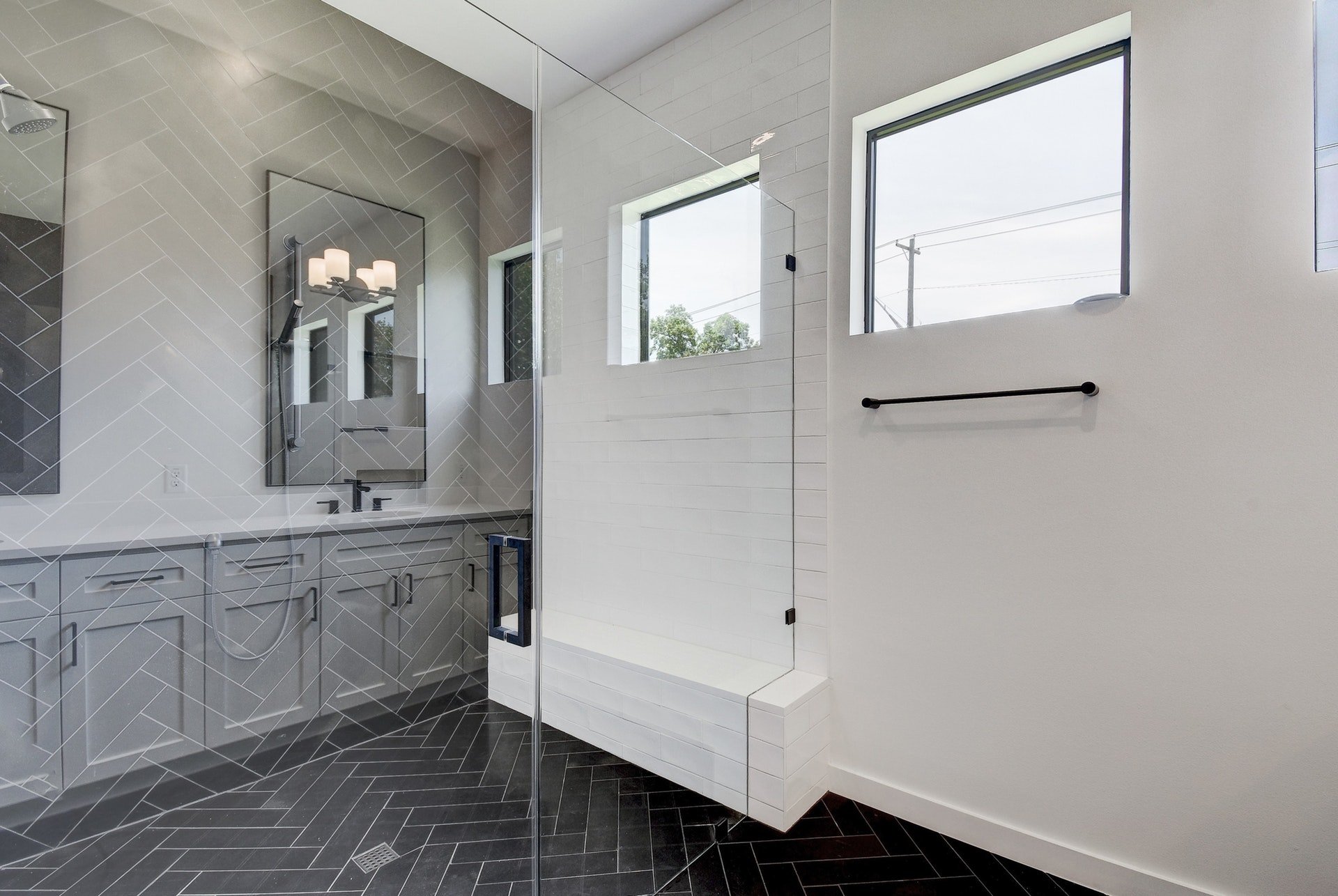 Modern bathroom with black herringbone tile flooring, a walk-in shower with a frameless glass door, white subway tile walls, and two windows.