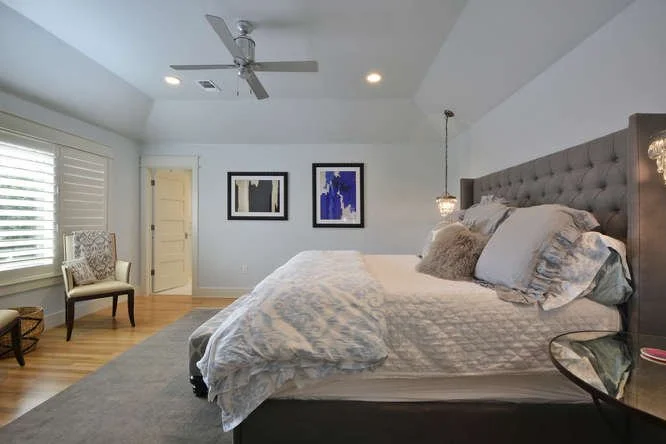 Bedroom with a large bed, gray upholstered headboard, and multiple pillows. Beside the bed are two hanging pendant lights. There's a gray area rug, a wooden chair with upholstery, and two framed abstract artworks on the wall. The room has white walls
