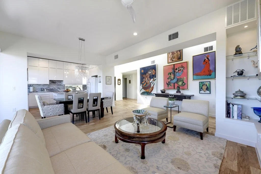 A bright, modern living room with beige sofa, white armchairs, a glass coffee table, and a gallery wall of colorful Asian-inspired artwork, adjacent to an open-concept kitchen with white cabinets and a marble backsplash.