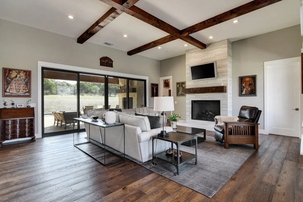 Living room with a beige couch, a wooden armchair, a black coffee table, a TV mounted above a fireplace, a patterned rug, and sliding glass doors leading outside to an outdoor dining area.