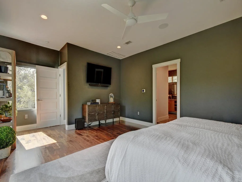 A bedroom with a partially visible bed with a white comforter, green walls, a ceiling fan, a wall-mounted TV, a wooden dresser, a door leading to a bathroom, a window with sunlight, and a wooden bookshelf with plants and decor.