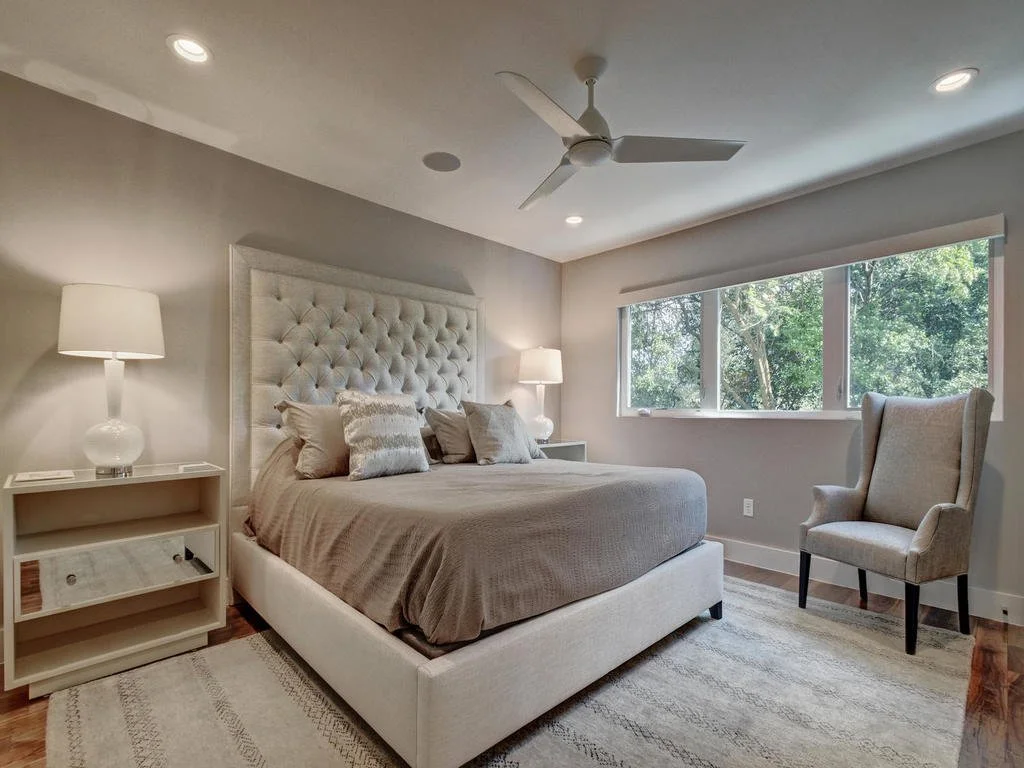 A bedroom with a large upholstered bed, nightstands with lamps, a beige armchair, a ceiling fan, large window, and hardwood floors.