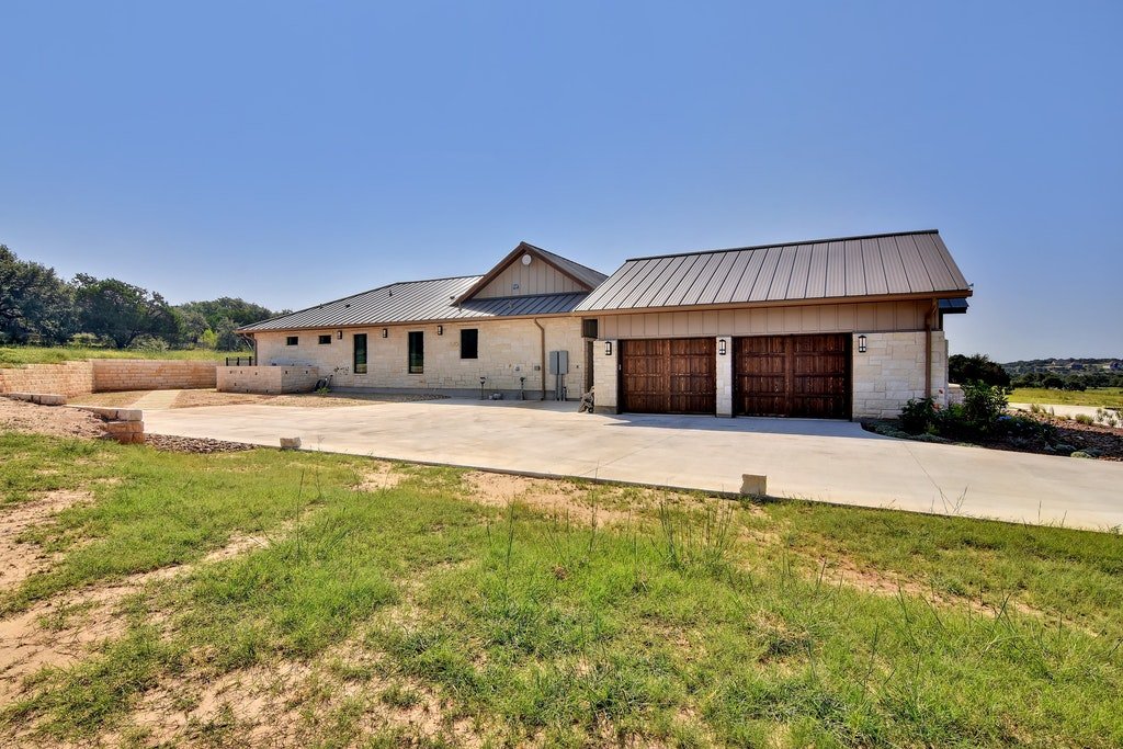 Newly constructed house with stone and wood exterior, metal roof, and two garage doors, situated on a concrete driveway with grassy front yard, under a clear blue sky.