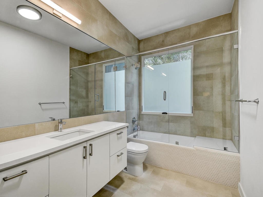 Modern bathroom with white vanity, large mirror, toilet, shower with glass doors, and beige tiled walls and floor.