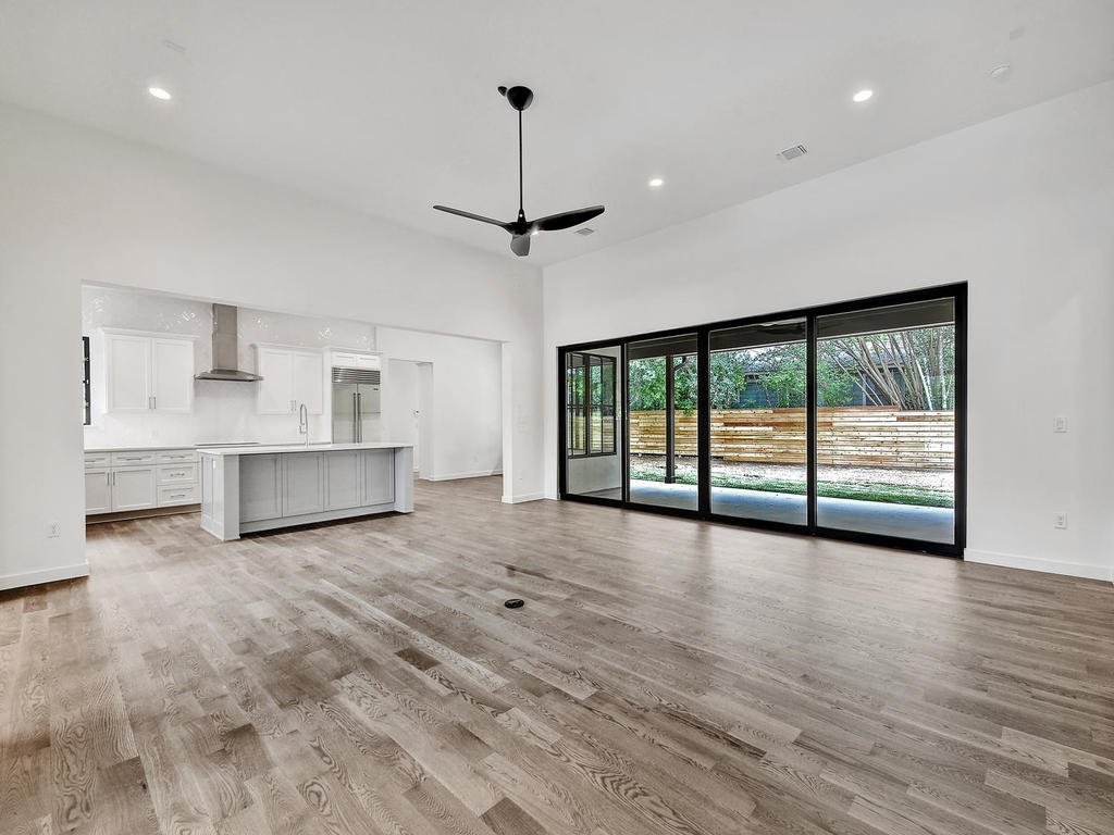 Empty living room with large glass sliding doors, ceiling fan, and modern kitchen in background.