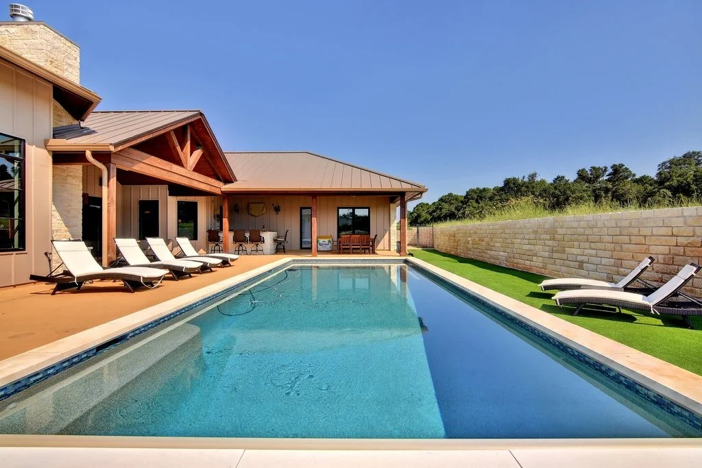 A backyard with a rectangular swimming pool, lounge chairs, a patio, and a house with a wooden porch and outdoor dining area under a clear blue sky.