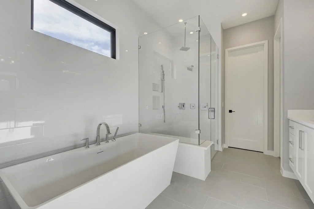 Modern bathroom with white bathtub, glass-enclosed shower, gray tile floor, white walls, and a window showing blue sky.