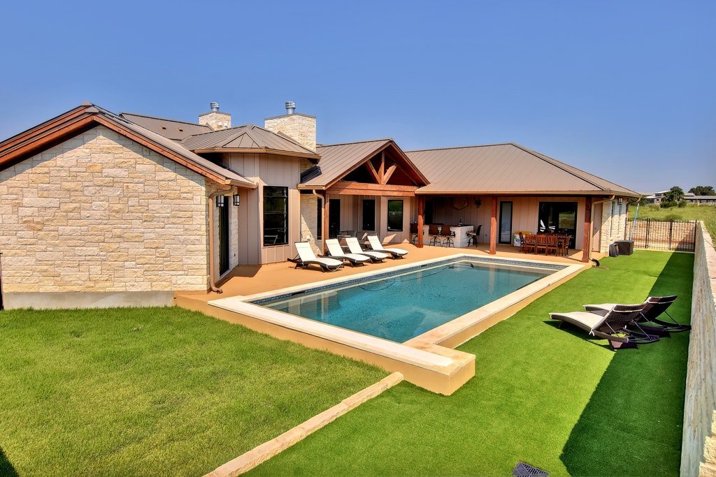 Backyard with a rectangular swimming pool, lounge chairs, and a patio with outdoor furniture, behind a house with stone and wood siding under a clear blue sky.